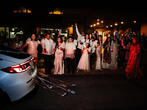 Large group of formally dressed guests gathered around a white car at night under warm outdoor lighting.
