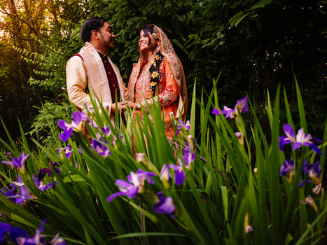 Couple in traditional wedding attire standing among purple flowers in a garden at golden hour.