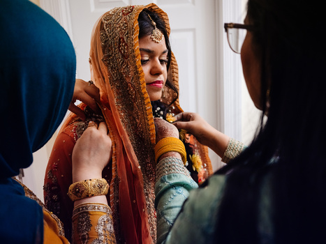 Woman in traditional gold and orange sari being dressed by two people during bridal preparation.