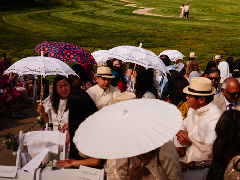 Crowd of people holding white and colorful umbrellas at an outdoor daytime event on a grassy lawn.