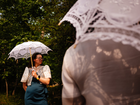 Woman holding decorative lace umbrella in sharp focus with blurred figure in background among green foliage.