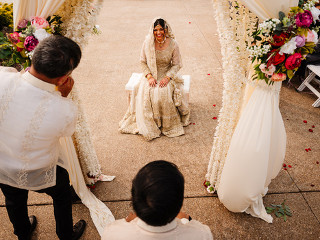 Aerial view of bride seated centrally while two grooms in white shirts arrange her bridal veil during wedding ceremony.