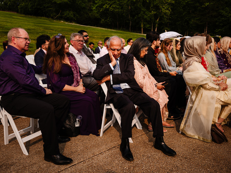 Guests seated outdoors at a formal event on a sunny day with green lawn and trees in background.