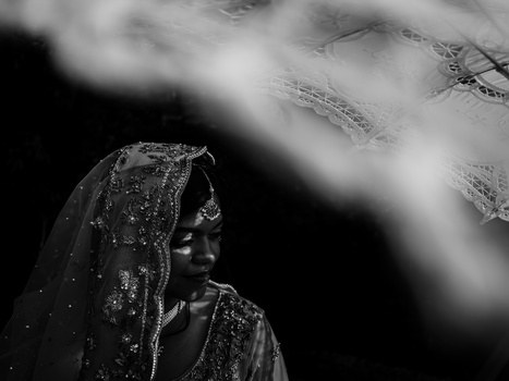 Woman wearing ornate traditional dress and headscarf, photographed in profile against a blurred mountainous background.