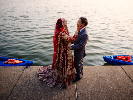 Couple in formal attire standing on a waterfront pier facing each other, with boats and calm water in the background.