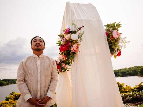 Man in cream-colored kurta standing before a draped white fabric backdrop decorated with pink and red roses beside a waterfront landscape.