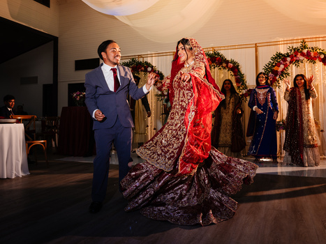 Groom in navy suit watches bride in red and gold lengha with dramatic embroidered train during wedding ceremony.