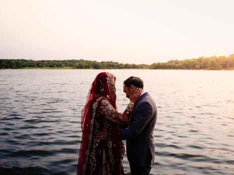 Couple in traditional red and dark clothing standing together by a calm river at golden hour.