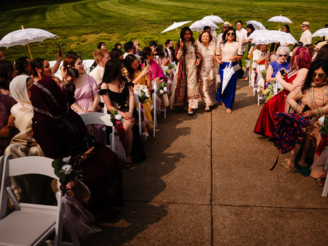 Outdoor wedding ceremony with guests seated under umbrellas on a tree-lined green lawn.