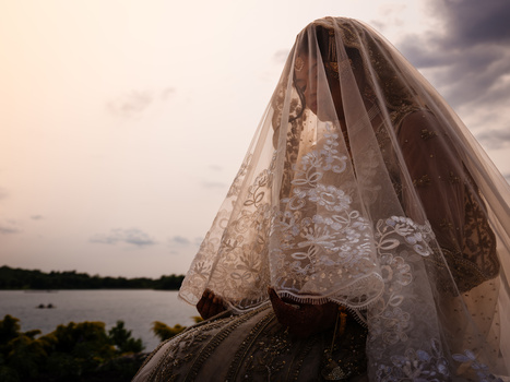 Bride wearing an ornate lace veil and beaded wedding dress, photographed from behind against a lakeside landscape at golden hour.