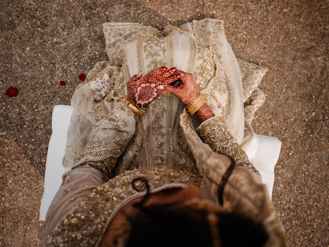 Overhead view of bride's hands adorned with henna and jewelry, holding red flower petals over ornate fabric.