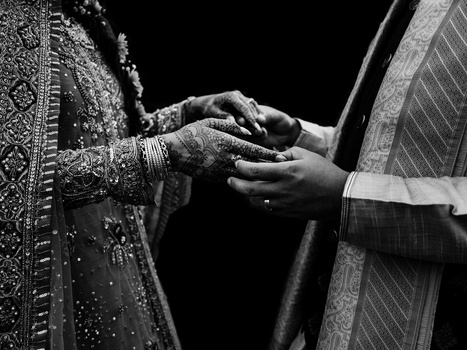 Hands of two people in ornate traditional clothing exchanging rings during a wedding ceremony against a black background.