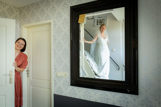 Woman in white dress reflected in large black-framed mirror on floral wallpapered wall.