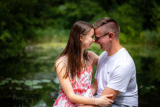 Couple facing each other by a pond with lush green foliage in the background.