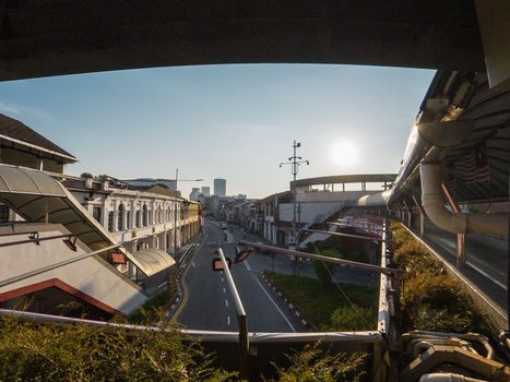 Urban cityscape photographed from an elevated covered structure, showing a street intersection with buildings and distant skyscrapers under clear sky.