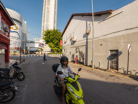 Motorcyclist in neon yellow jacket riding through urban street with tall building and residential structures.