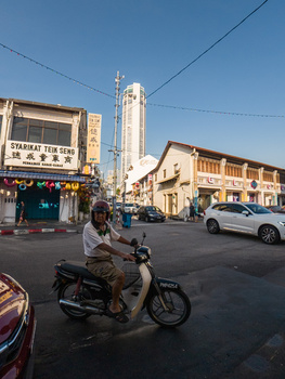 Man on a motorcycle riding through a busy urban street with historic storefronts and a tall modern building in the background.