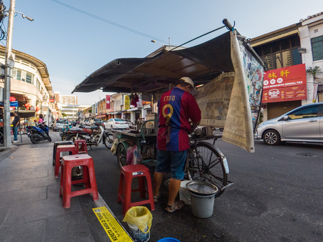Street vendor with red plastic stools operates a food stall beneath a canvas awning on an urban sidewalk.