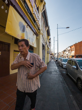 Man in striped shirt standing on a residential street lined with parked cars and yellow buildings.