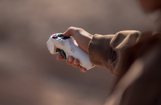 Hand holding a white drone controller with orange accents and visible buttons against a blurred background.
