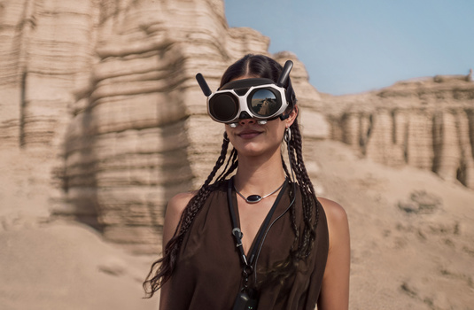 Person wearing oversized steampunk-style goggles and dark clothing against desert rock formations.