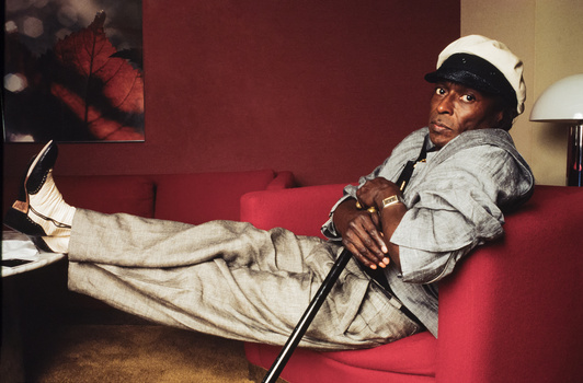 Man in white cap and gray jacket reclines on red couch while holding a golf club.