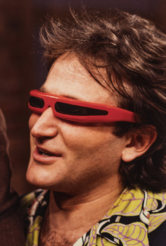 Man wearing red-framed sunglasses and yellow patterned shirt against dark background.