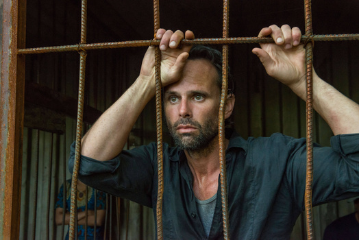 Bearded man gripping rope in front of a barred window, direct gaze toward camera.