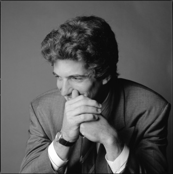 Man in suit with hand to face in contemplative pose against neutral background.