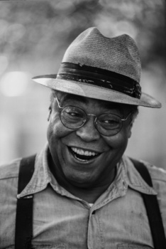 Man wearing fedora and glasses smiling broadly at camera in black and white portrait.