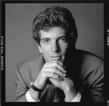 Black and white portrait of a man with dark wavy hair, chin resting on clasped hands, wearing a collared shirt.