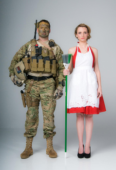 Military personnel in camouflage uniform standing next to person in white and red dress against neutral background.
