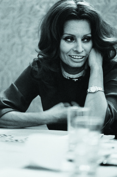 Woman in dark clothing smiling at camera with hand near face, posed at table in black and white portrait.