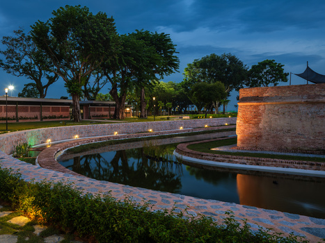 Curved waterway at dusk with illuminated pathway, trees, and brick fortification wall reflected in still water.