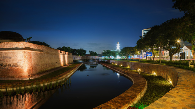 Illuminated fortress wall and canal at dusk with historic buildings reflected in still water.