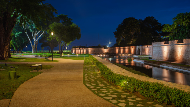 Illuminated fortress wall reflected in canal at twilight, with manicured lawn and trees in foreground.