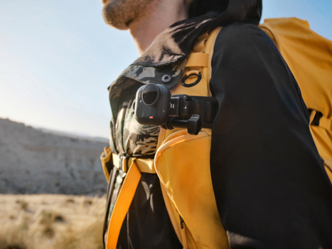 Photographer wearing a yellow backpack carries a compact camera on chest strap while hiking in desert terrain.