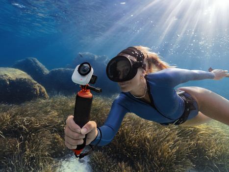 Snorkeler in blue wetsuit holding underwater camera housing, photographing seagrass on shallow ocean floor with sunlight filtering through water.