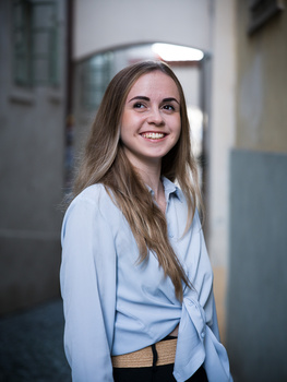 Young woman with long brown hair smiling at camera in well-lit indoor hallway.