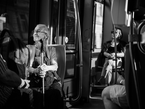 Elderly man with glasses sitting on public transit, holding onto a pole while looking upward.
