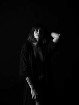 Woman in dark clothing illuminated by dramatic side lighting against a black background.