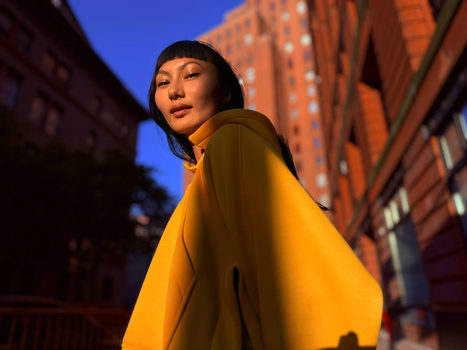 Woman in yellow coat turning toward camera on urban street at dusk with warm building lights in background.