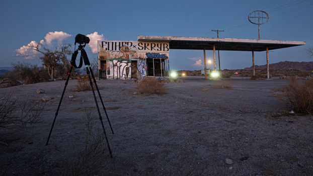 Tripod-mounted camera on desert ground facing an abandoned gas station at dusk.