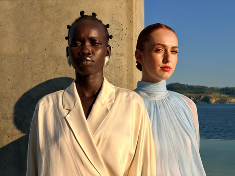 Two people in light-colored clothing positioned against a concrete wall with a coastal waterscape behind them.