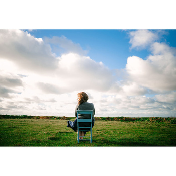 Person sitting on a chair in an open field, facing away toward the horizon under a partly cloudy sky.