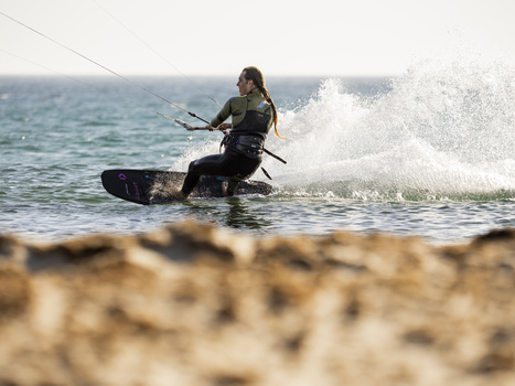 Kitesurfer riding a wave in shallow water near a sandy beach with a large kite line visible overhead.