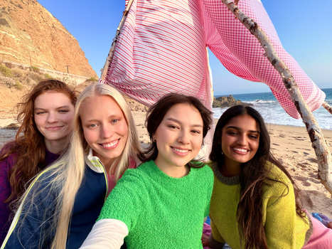 Four young women smiling at camera on beach with pink and white fabric structure and coastal cliffs behind them.