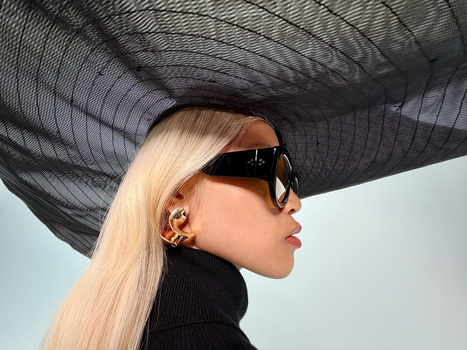 Blonde woman wearing oversized black sunglasses and a black turtleneck, positioned in profile against a clear sky with a large black mesh hat overhead.
