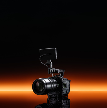 Professional cinema camera with telephoto lens mounted on tripod against warm orange and black background.
