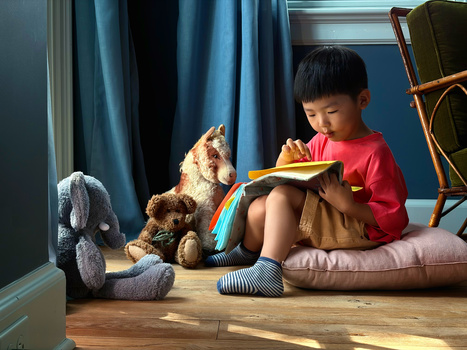Young child in red shirt reading a book while seated on floor surrounded by stuffed animals.